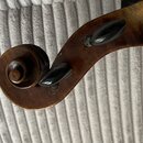 Detail hlavy smyčcového nástroje v hnědé barvě s výraznou spirálovitou volutou. Jsou vidět dva černé ladicí kolíky. Povrch vykazuje známky opotřebení…