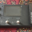 Černý kytarový procesor s displejem a expresním pedálem. Na přístroji jsou tři spínací tlačítka s nápisy - STOMP/RIG, - LOOPER a - TUNER oraz nápis H…