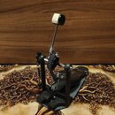 Černý bicí pedál s kovovou konstrukcí a bílou pumpliškou na konci šlapáku. Mechanismus pohonu je řešen pomocí ozubeného řemenu. Na nástroji nejsou žá…