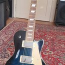 Tmavě modrá elektrická kytara s tvarem singlecut a bílým pickguardem. Na hlavě je viditelné logo Gibson. Kytara má dva humbuckery s kovovými kryty, č…