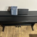 Černý digitální piano v dřevěněm korpusu s trojpedálem. Na nástroji je nápis YAMAHA a vedle něj stojí manuál s textem ARIUS, YDP-164, YDP-144 a logem…
