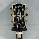 Hlava kytary značky Epiphone s bílým lemováním a černým povrchem. Uprostřed je umístěna vložka s geometrickým vzorem, ladicí mechanismy jsou zlaté ba…