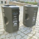 Dvě černé plastové aktivní reproduktory stojící na dlažební kostce. Na zadním panelu obou kusů je viditelné logo s hlavou vlka a nápis TX-AMP12. Jsou…