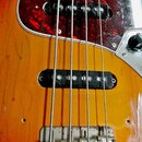Detail elektrické basové kytary s tělem v žlutě-oranžovém přechodovém lakování. Nástroj má červený torties pickguard a dva černé snímače. Povrch laku…