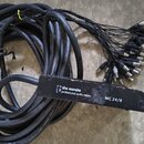 Černý svazek audio kabelů s kovovými konektory. Na černé držáku je bílý nápis „the ssnnake professional audio cables“ a níže označení „MC 24/8“. Povr…