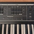Detailní pohled na ovládací panel černého digitálního klavíru. Panel obsahuje sekce s nápisy PIANO, SYNTH a DRUM, které jsou vybaveny různými tlačítk…