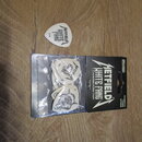 Bílé kytarové trsátka v plastovém obalu a jedno volné trsátko. Na trsátkách je černý nápis HETFIELD WHITE FANG a číslo 1.74. Obal je černý s bílým ná…