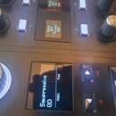 Detailní záběr na ovládací panel černého elektronického syntetizátoru s několika otočnými knoflíky a stříbrnými tlačítky. Na horním displeji jsou vid…