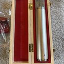 Stříbrný kondenzátorový mikrofon v dřevěném kufru s červeným vnitřním polstrováním. Na vnitřní straně víka je nápis FLEA CUSTOM MADE MICROPHONES. Spo…