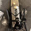 Stříbrný kondenzátorový mikrofon v černém pavoukovém držáku. Na těle mikrofonu je vidět displej s nápisy 8, -80, -40 a -10. Nad displejem svítí malá…