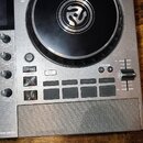 Stříbrný DJ 컨트롤러 s nápisem MIXSTREAM PRO GO. V horní části je velký černý jog-wheel s bílým symbolem uprostřed. Zařízení obsahuje různé ovládací knof…