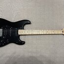 Černá elektrická kytara s jasným dřevěným hmatníkem a černým pickguardem. Na hlavě je viditelné logo Squier a nápis Stratocaster. Nástroj vykazuje zn…