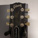 Záběr na hlavu černé elektrické kytary s bílými ladicími kolíky. Na hlavě jsou viditelné nápisy „Gibson“, „Les Paul MODEL“ a na krytce v dolní části…