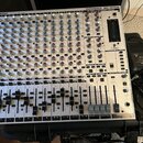 Stříbrný mixážní pult s nápisem EURORACK MX 2642A a BEHRINGER. Zařízení obsahuje řadu modrých a červených otočných knoflíků, posuvníky a tlačítka. Na…