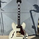 Kytara typu semi-akustika v krémové barvě s hnědočerveným pickguardem a zlatými komponenty. Na hlavě je bílý nápis Epiphone. Hриф má vložky z perlece…