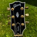 Hlava kytary v černé barvě s bílým lemováním a zlatými ladičkami. V horní části je nápis Gibson, v dolní části na krytu ladicího mechanismu nápis Cus…