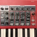 Červený digitální klavír s ovládací sekcí obsahující knoflíky, tlačítka a displej. Na panelu jsou viditelné nápisy PROGRAM, SAMPLE SYNTH a EFFECTS s…