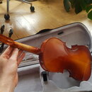 Hnědá lakovaná housle v otevřeném šedém pouzdře. Nástroj má klasický tvar a hladký povrch bez viditelných značek či nápisů. Na hlavě jsou černé kolík…