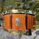 Dřevěný buben s přirozeným povrchem a zlatými kovovými obručemi. Uprostřed korpusu je kovový štítek s logem a nápisem CUSTOM DRUMS. Nástroj vykazuje…