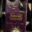 Fialový kytarový efekt s nápisy MXR CUSTOM SHOP a DUKE of TONE. Na horní straně jsou tři černé otočné knoflíky a přepínač s označením OD, BOOST, DIST…