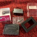 Na červeném povrchu je položen kytarový snímač typu humbucker v kovovém krytu s černými vodiči. Vedle něj je bílá krabička a červený papírový obal s…