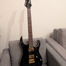 Černá elektrická kytara s tmavým hnedým hmatníkem a zlatým hardwarem. Na hlavě je bílý nápis Ibanez. Kytara má dva humbuckery, dvě otočné potenciomet…