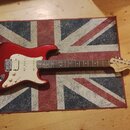 Červená elektrická kytara s bílým pickguardem a hnědým hmatníkem. Na hlavě je viditelné logo Squier STRATOCASTER. Nástroj leží na koberci s motivem b…