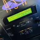 Detailní záběr na ovládací panel černého elektronického zařízení. Zelený LCD displej zobrazuje text: „L01 SYNOTHER: Sync' 0' Goob ROM09:028“. Pod dis…