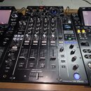 Černý DJ mixér s nápisem PROFESSIONAL MIXER DJM-900NXS2 a logem Pioneer DJ. Zařízení obsahuje početné posuvníky, otočné knoflíky a tlačítka v různých…