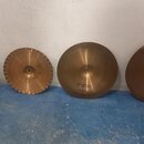 Čtyři mosazné činely různých velikostí opřené o bílou zeď na modré podlaze. Na dvou pravých činelech je viditelný nápis PAiste. U levých dvou činelů…