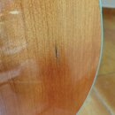 Detailní záběr na lakovaný dřevěný povrch akustického nástroje v hnědavé barvě. Uprostřed snímku je vidět svislá škrábance v laku a dřevě. Žádné znač…