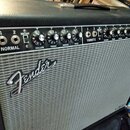 Kytarový zesilovač s černým povrchem a stříbrnou síťovkou. Na předním panelu je nápis Fender a ovládací prvky s popisky NORMAL, BRIGHT, VOLUME, TREBL…
