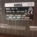 Fotografie detailu štítku na zadní straně přístroje. Na černém štítku je bílým textem uveden výrobce KORG, MODEL GS1-73 a SERIAL No. 001458. Je zde u…