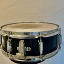 Černý snare drum s kovovými obručmi a mechanismem na napnutí blány. Povrch blány je viditelně znečištěný a opotřebovaný, kovové části vykazují známky…