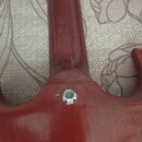 Detail zadní strany kytary s červeným lakem a viditelnou strukturou dřeva. V místě spoje krku s tělem je umístěn kovový šroub s maticí. Povrch laku v…