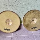 Dvě mosazné činely opřené o světlou stěnu. Oba mají v dolní části černý nápis pAiste a pod ním menší text MADE IN SWITZERLAND. Levý činel má hladký p…