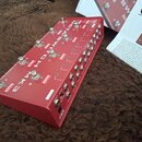 Červený kovový pedalboard s nápisem OCTA-SWITCH MK3. Na horní straně se nachází osm stříbrných spínacích tlačítek, vedle nich jsou bílé nápisy s čísl…