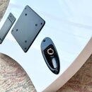 Detail zadní strany bílé kytary s černými plastovými kryty. Na fotce je vidět čtvercový kryt elektroniky a oválný kryt s výstupní zásuvkou, oba připe…
