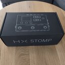 Černá papírová krabice s bílým lineárním nákresem efektového pedálu. Na přední straně je bílý nápis HX STOMP. Krabice vykazuje mírné opotřebení hran.