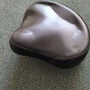 Černý polstrovaný sedák v ergonomickém tvaru. Povrch je z hladkého materiálu s viditelným prošíváním po okraji. Na fotce nejsou žádné viditelné značk…