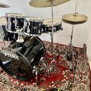 Černá bubnová sada značky TAMA s kovovými mechanismy a dvěma činely na stojanách. Sada obsahuje basový buben s bílým nápisem TAMA, dva tom-tomy a jed…