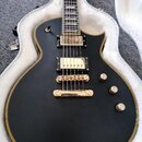 Černá elektrická kytara s jednosřezovým tělem a zlatým lemováním, položená v bílém tvrdém kufru. Nástroj má dva humbuckery, most a strunovník v zlaté…