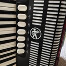 Detail akordeonu s klávesovou klaviaturou a černým plastovým krytem. Na krytu je umístěno kulaté bílé logo s nápisem HOHNER a siluetou postavy. Vedle…