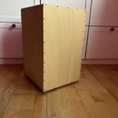 Světle hnědý, dřevěný reproduktorový box s hranatým tvarem. Boční stěna je spojena šrouby viditelnými v řadě podél hran. Povrch dřeva je hladký bez v…