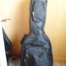 Černý polstrovatý pouzdro (gig bag) na hudební nástroj. Má dvě zipové kapsy, jednu úzkou v horní části a jednu širší ve spodní části. Povrch materiál…