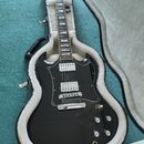 Černá elektrická kytara s dvojstupňovými humbuckery a bílým pickguardem, umístěná v otevřeném černém kufru s bílou vnitřní výstylou. Na hlavě nástroj…