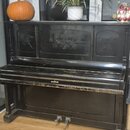 Černý vzpravený piano v lakovaném provedení s dekorativními rytinami na horním panelu. Povrch vykazuje viditelné opotřebení, zejména odřesy a škrában…