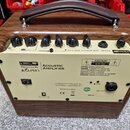 Akustický zesilovač v dřevěném šasi s hnědým koženým úchem. Na horní straně jsou ovládací knoflíky a nápisy Bluetooth, FX a KINSMAN 25W ACOUSTIC AMPL…