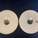 Dvě zlatavé činely typu hi-haty s hloubkovým rýhováním povrchu. Na pravém číneli je černý nápis AAX Medium Hats 14"/35 cm. Oba činely jsou v čistém s…