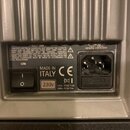 Detail zadní strany zařízení s hliníkovým chladičem. Je zde vypínač, napájecí konektor a štítky s nápisy: CAUTION, MADE IN ITALY, CE, 230V a údaje o…